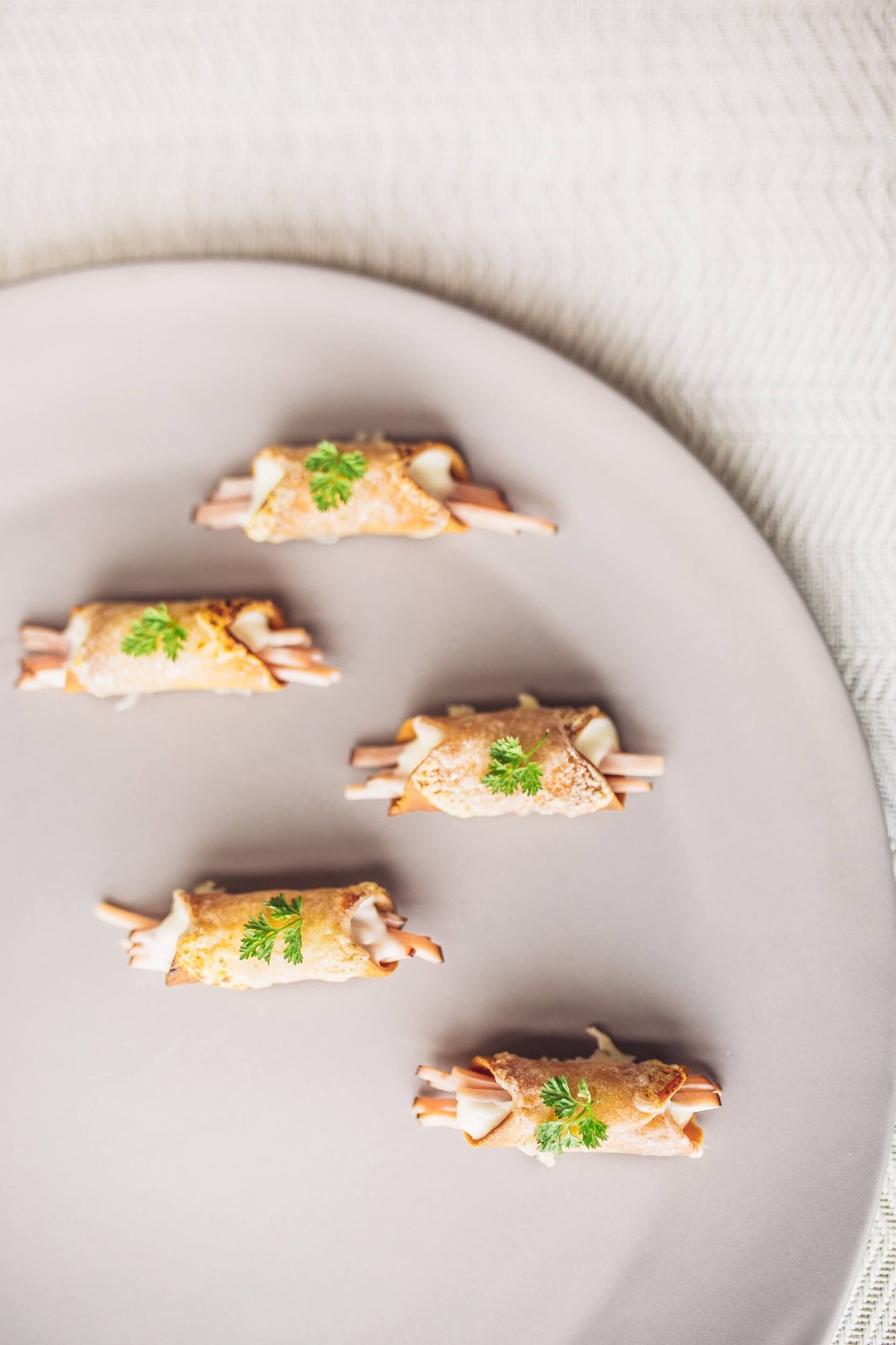 Five golden-brown savory crepe rolls filled with sliced deli meat and garnished with fresh parsley on a round gray plate