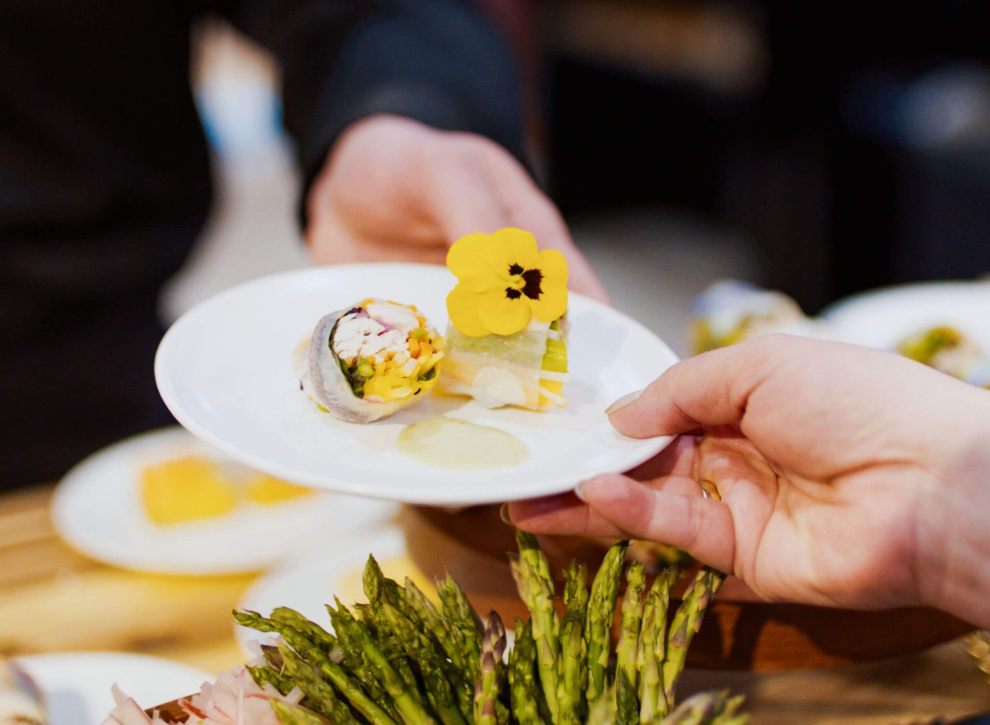  Chef's hands presenting a white plate with a fresh spring roll, edible yellow pansy flower, and citrus vinaigrette sauce, with fresh asparagus in the foreground