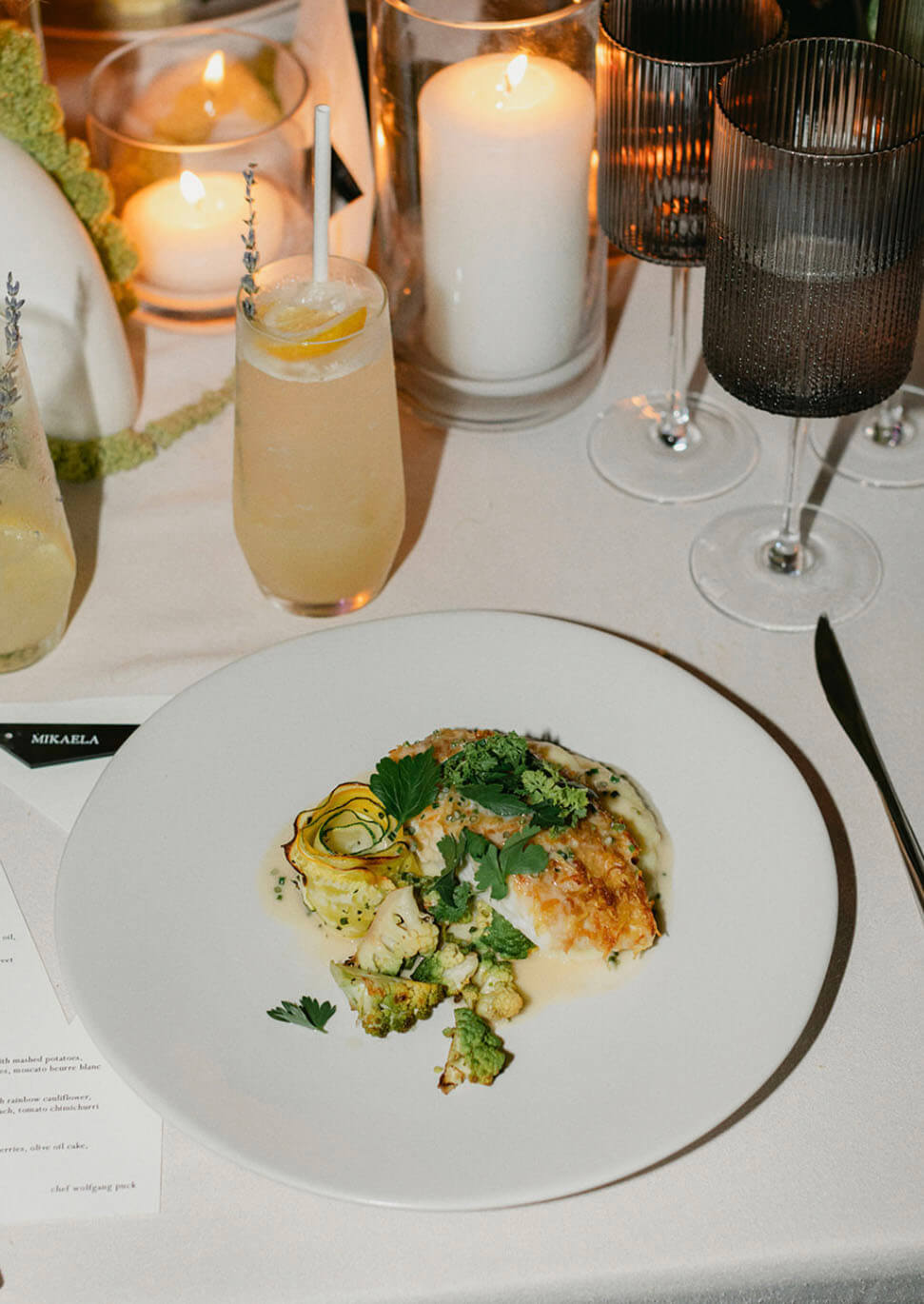 Elegant wedding dinner place setting featuring a pan-seared fish entrée with rainbow cauliflower and herb butter sauce, a signature cocktail, and a personalized place card