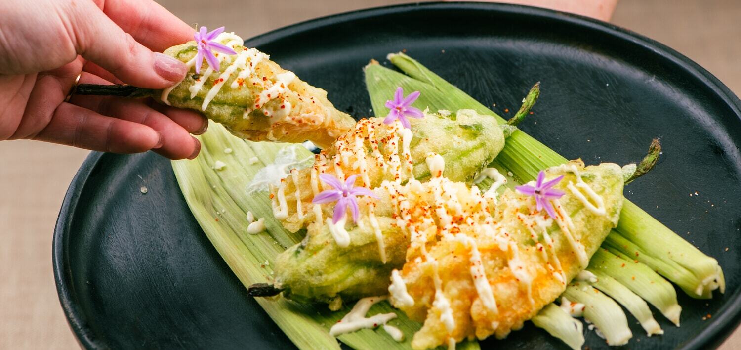 Crispy stuffed squash blossoms topped with creamy sauce, spices, and edible flowers on a black plate.