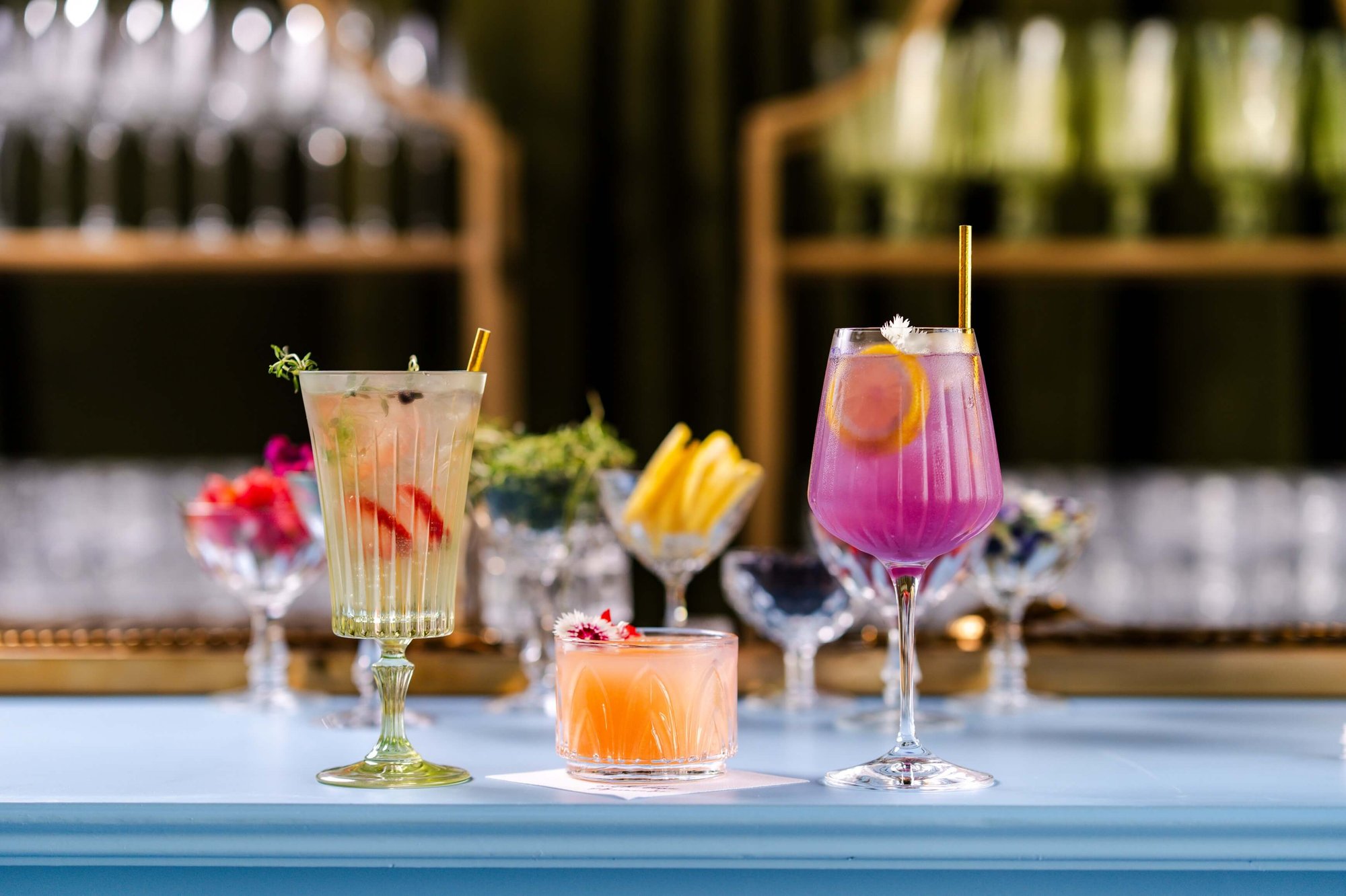 Three colorful craft cocktails displayed on a bar: a tall white herb cocktail, a peach cocktail in a rocks glass with an edible flower, and a vibrant purple cocktail in a wine glass with an orange slice.