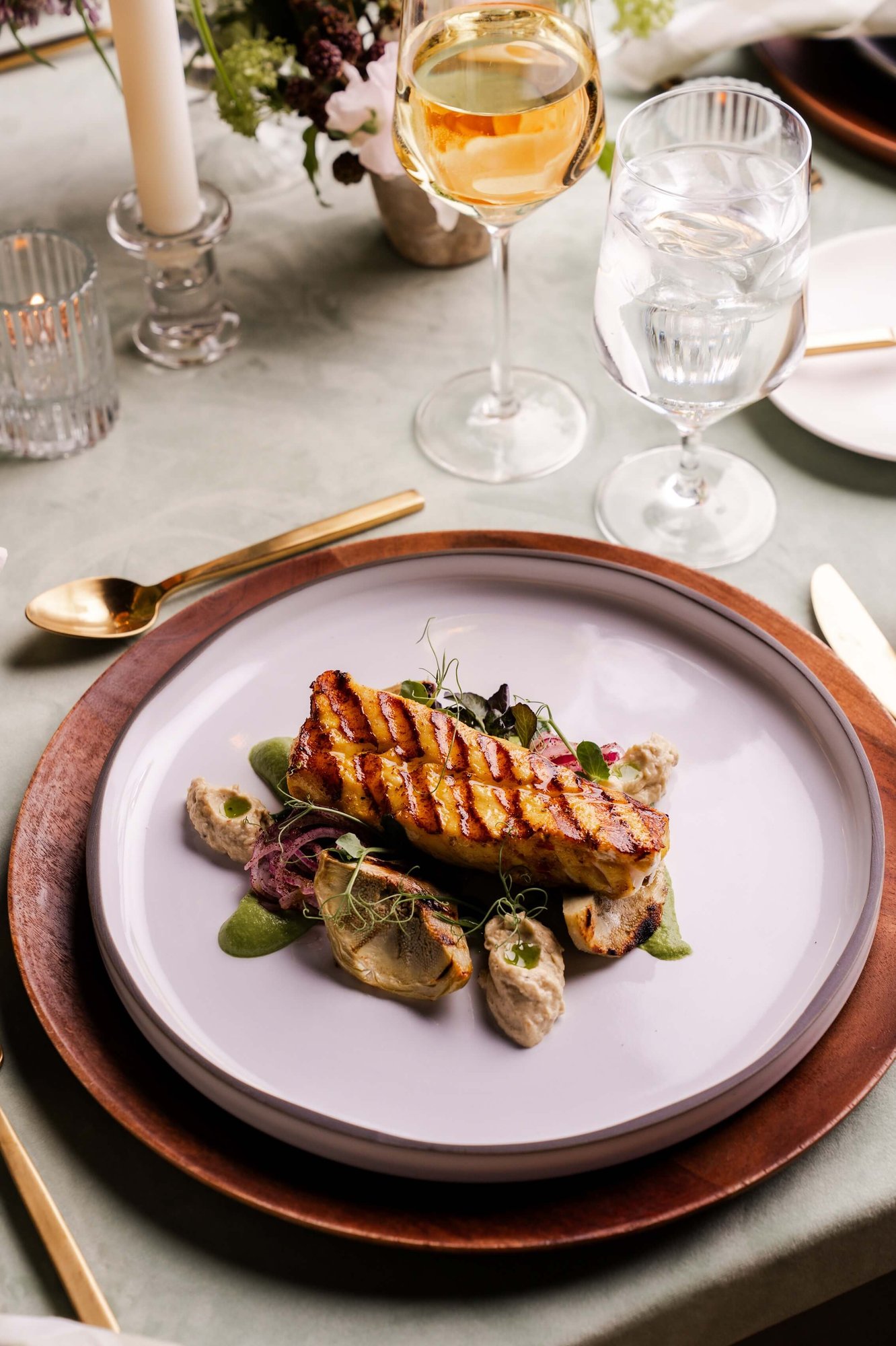 Grilled salmon fillet with artichoke, green herb purée, and microgreens on a lavender plate, with wine glasses and floral arrangements at an elegant dinner table