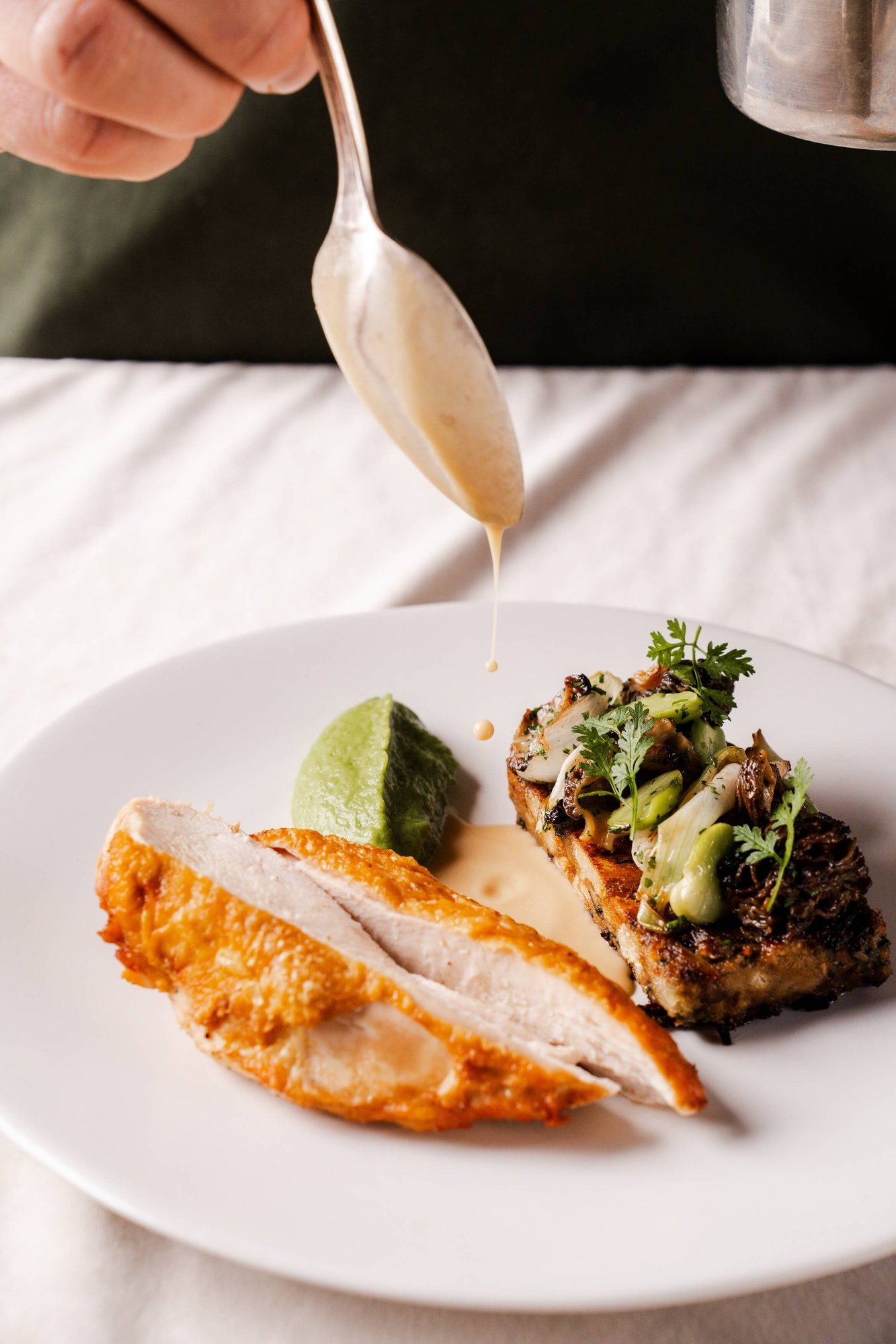 Grilled salmon fillet with artichoke, green herb purée, and microgreens on a lavender plate, with wine glasses and floral arrangements at an elegant dinner table