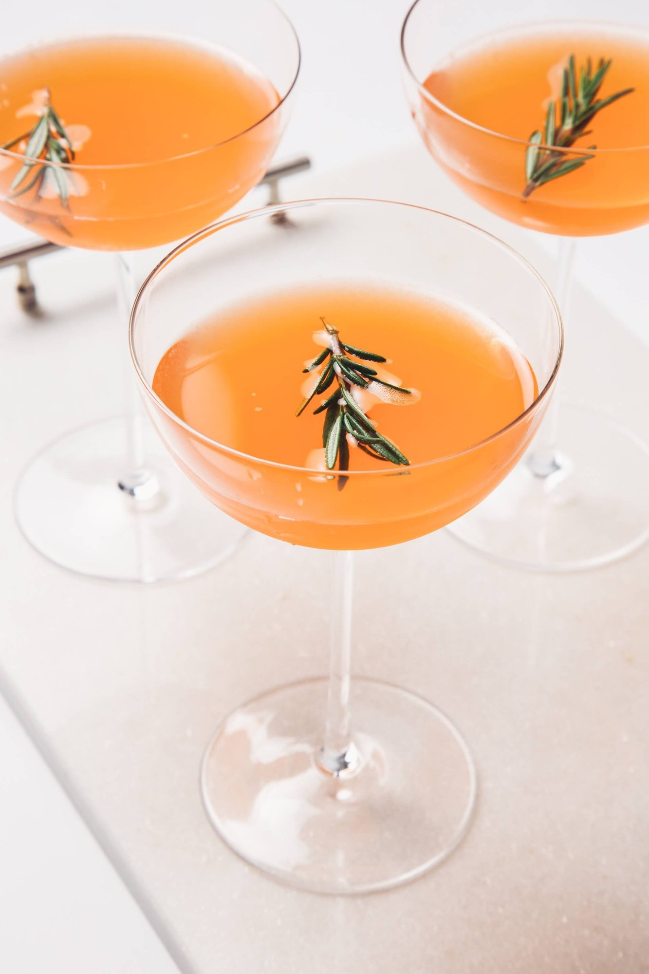 Three amber-orange cocktails in coupe glasses garnished with fresh rosemary sprigs, arranged on a white marble surface