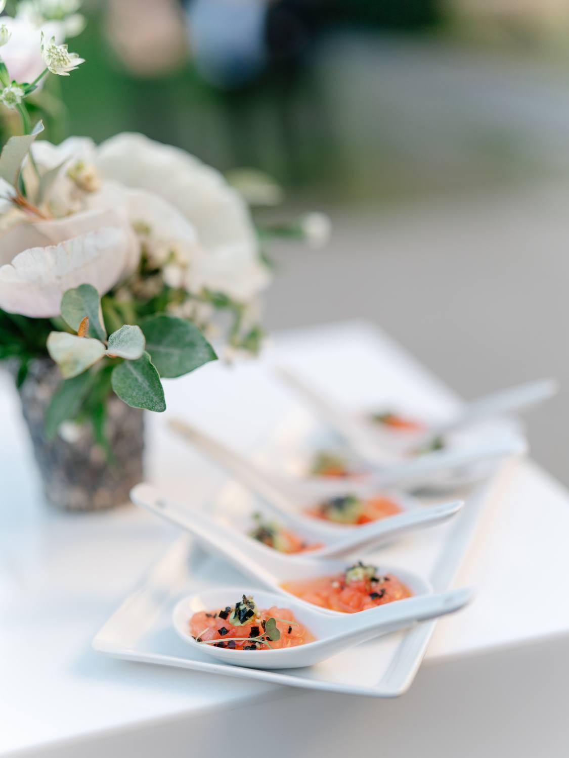 WPC Wedding Catering Elegant salmon tartare hors d'oeuvres served in white porcelain tasting spoons on a catering display tray alongside a white floral arrangement