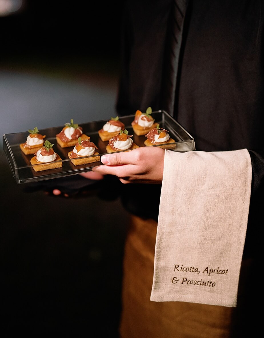 Uniformed server holding a tray of ricotta, apricot, and prosciutto crostini bites, with the dish name elegantly labeled on a white napkin