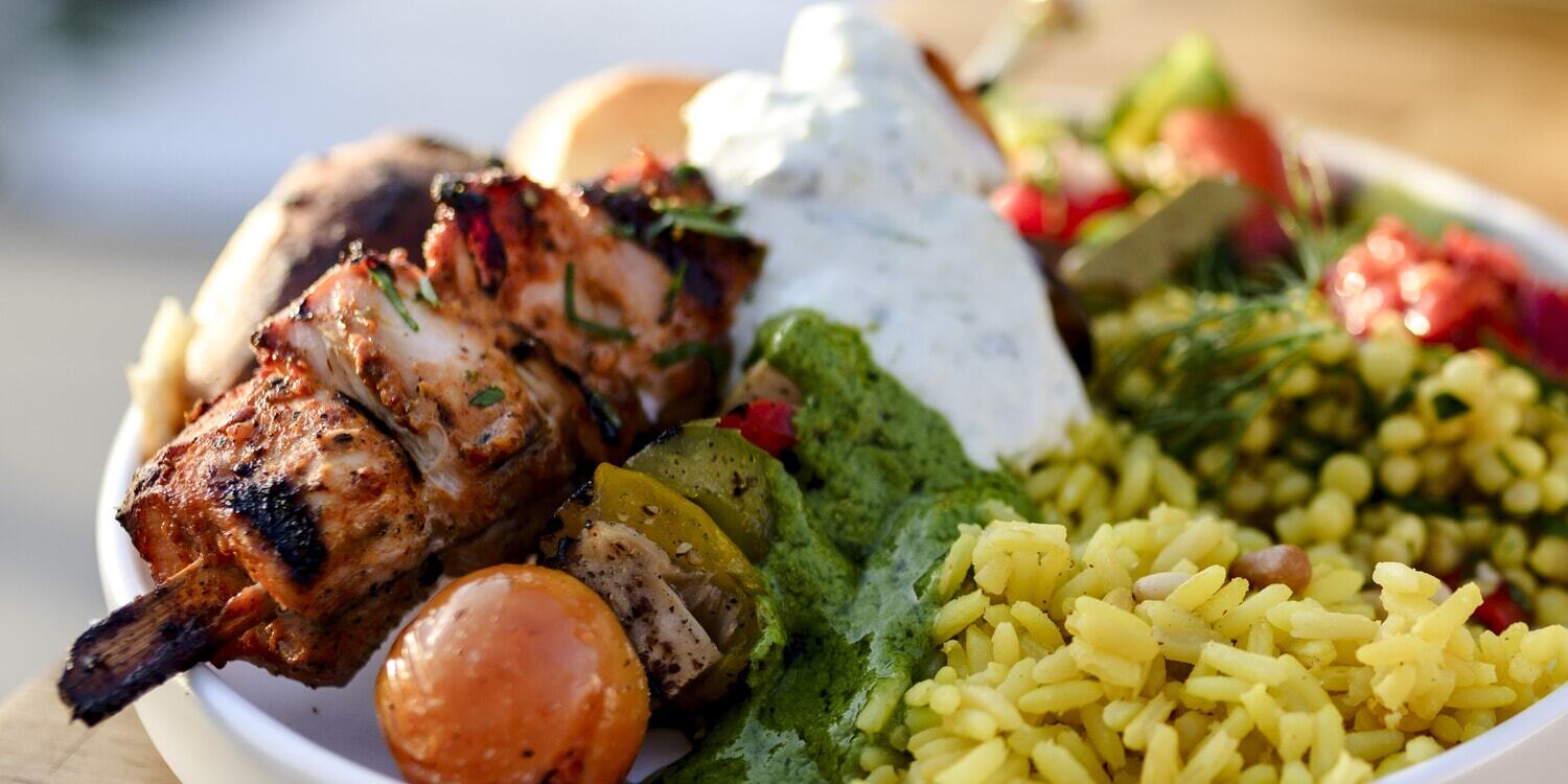 Grilled chicken skewers served with yellow rice, creamy herb sauce, grilled vegetables, and fresh salad.