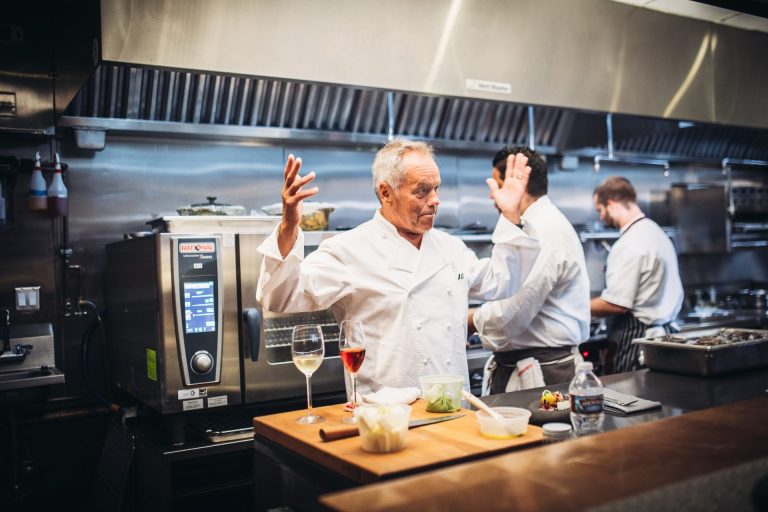 Wolfgang Puck standing in a kitchen with his hand up